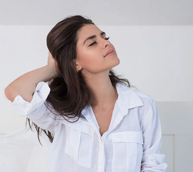 A woman with her eyes closed, enjoying a moment of calm, gently running her hand through her hair. She is wearing a relaxed white button-down shirt - B-12 at Truffles Aesthetics