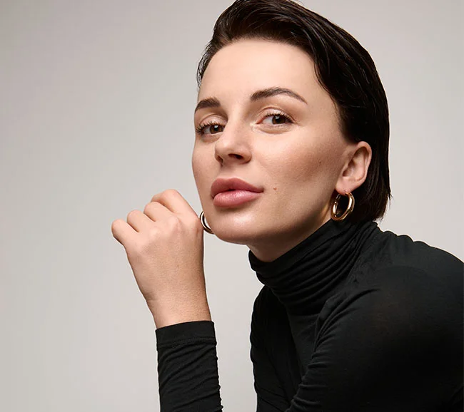 A confident woman with short dark hair wearing gold hoop earrings and a black turtleneck, leaning forward with a poised expression - Hydrafacial at Truffles Aesthetics