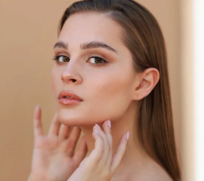 A close-up of a brunette woman touching her jawline with both hands, highlighting her smooth skin and subtle makeup with a neutral beige background - Kybella at Truffles Aesthetics