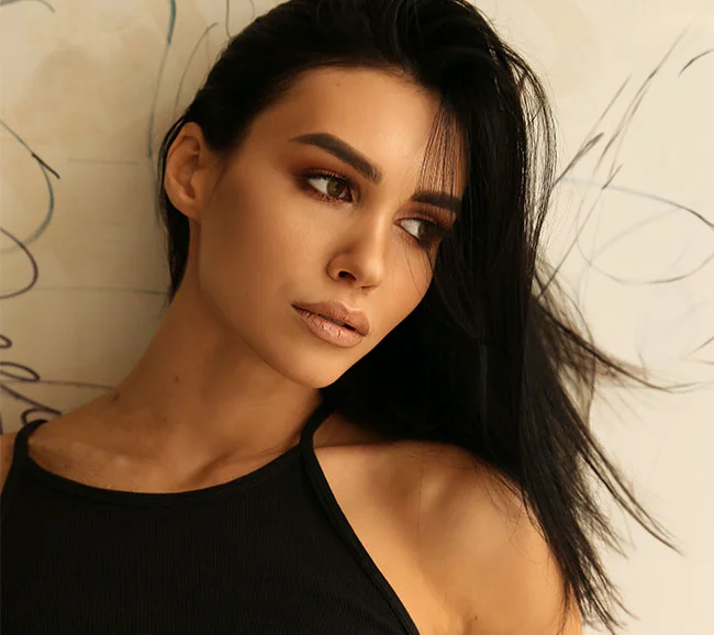 A woman with straight black hair and a black tank top gazes thoughtfully off to the side, with a soft, neutral background behind her - Masseters TMJ Treatment at Truffles Aesthetics